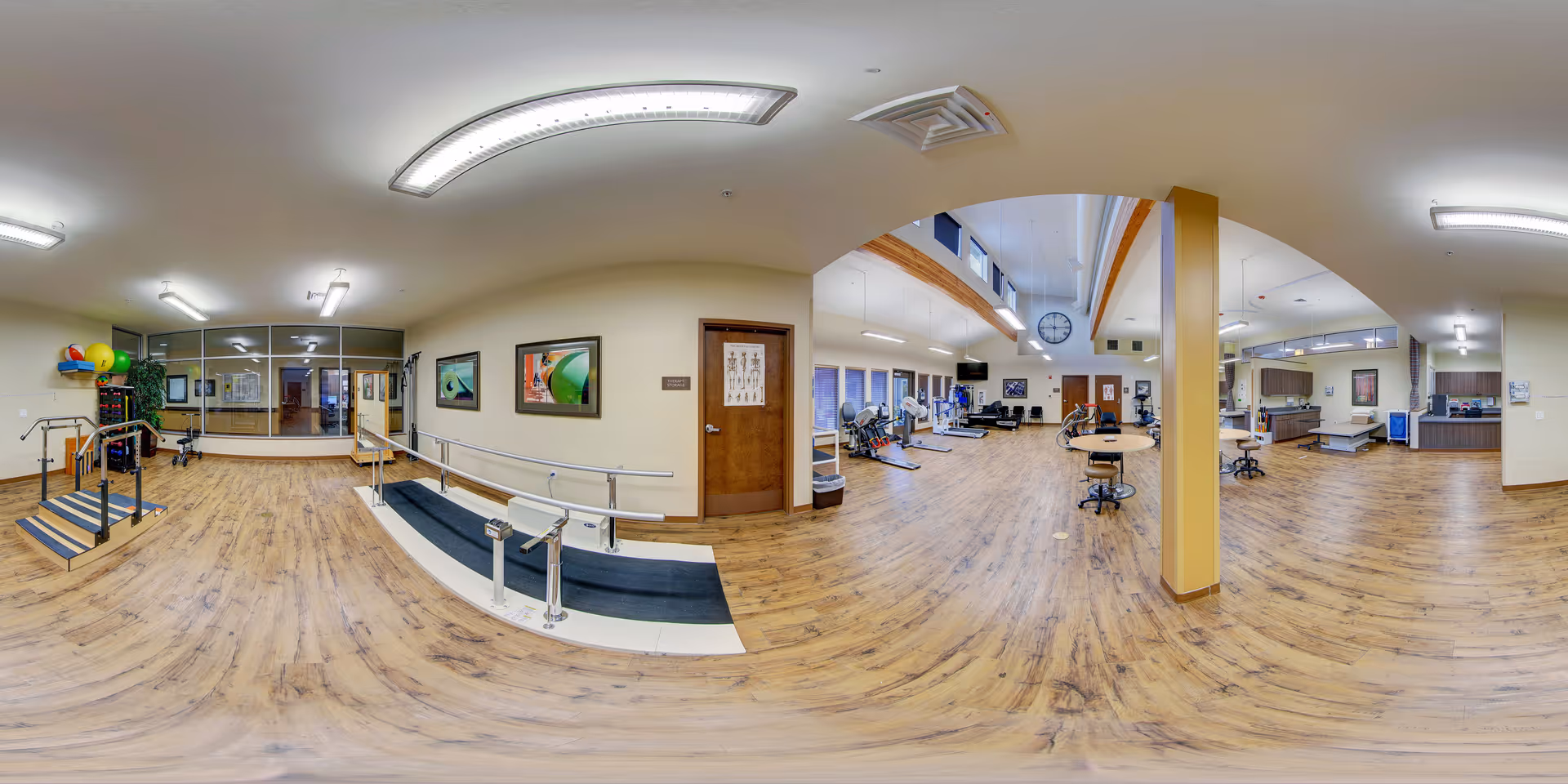 A spacious rehabilitation or physical therapy room with wooden flooring, parallel bars, a ramp, exercise equipment, therapy tables, and chairs. The room is well-lit with ceiling lights and has large windows along one wall. There are framed pictures on the walls and a clock hanging from the ceiling.