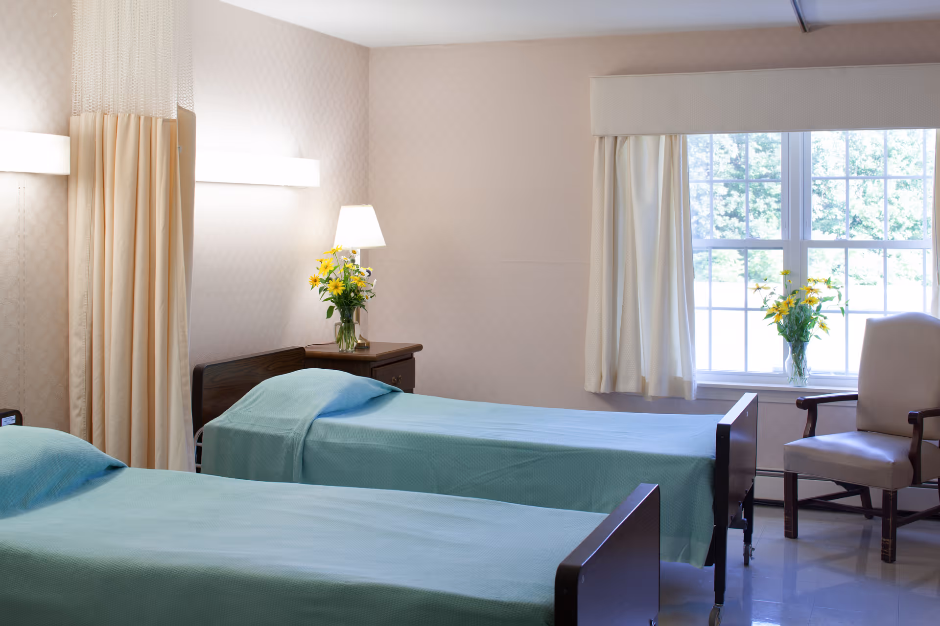 Two neatly made twin beds with teal covers in a bright senior room with a window, chair, nightstand and flowers.