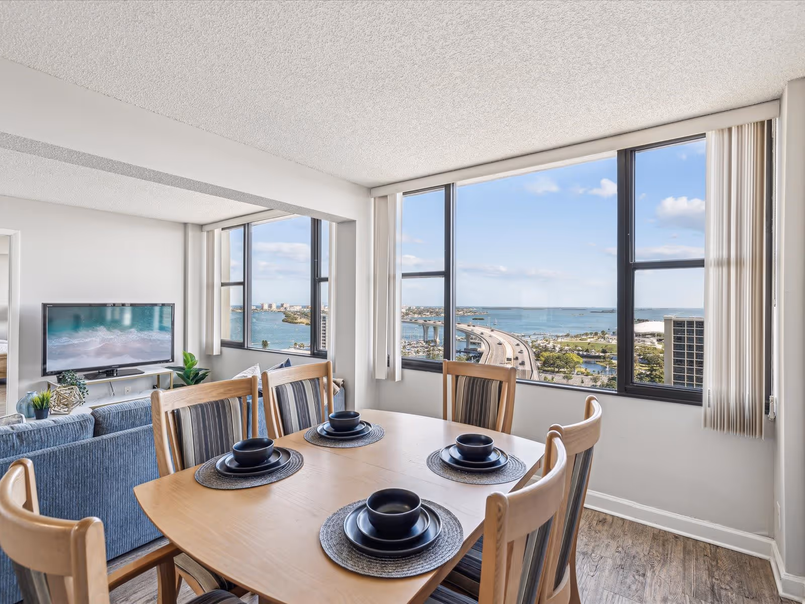 Dining area with a wooden table set for four with black bowls and plates on woven placemats, adjacent to a living room with a blue sofa and a TV showing a beach scene. Large windows offer a scenic view of a bridge, water, and cityscape under a blue sky with some clouds.