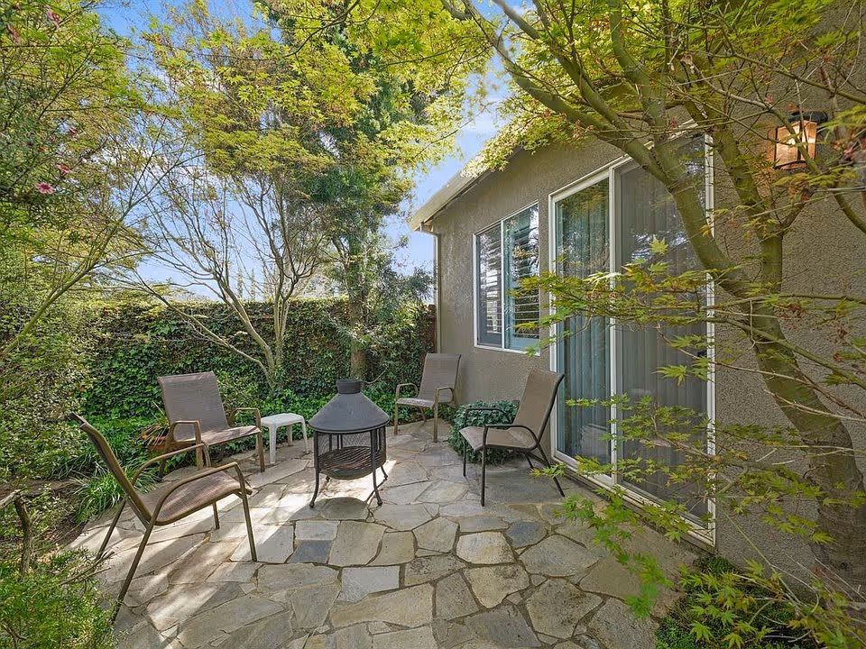 Stone patio beside a house with several outdoor chairs and a chiminea fire pit surrounded by trees and greenery.