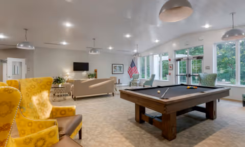 Bright communal lounge with a pool table, seating areas, large windows, and an American flag.