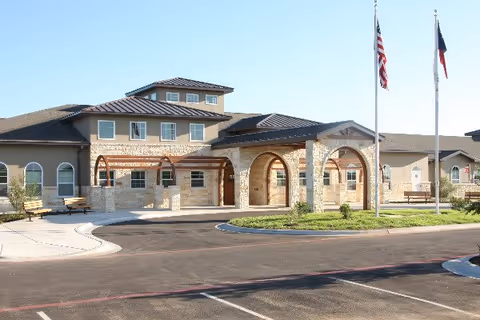 Front exterior of a senior living facility with an arched stone entrance, flagpoles, and a parking area.