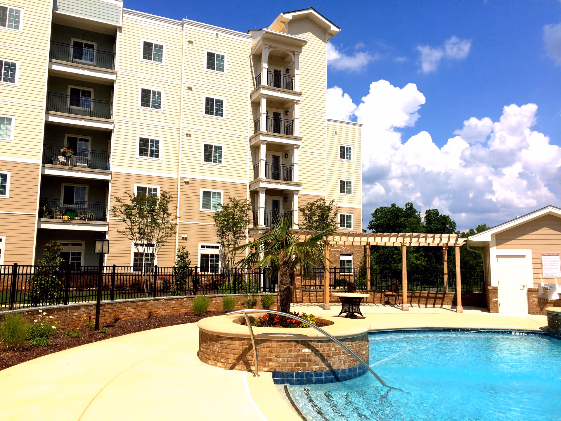 Outdoor view of a senior living facility with a swimming pool in the foreground, a small palm tree planted in a circular brick planter, a pergola with seating area, and a multi-story residential building in the background under a blue sky with scattered clouds.