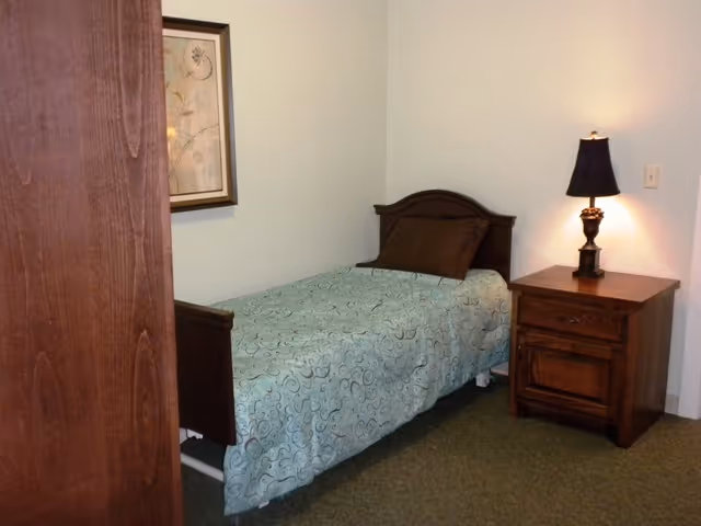 A small bedroom with a single bed covered with a light blue patterned bedspread and a brown pillow. Next to the bed is a wooden nightstand with a decorative lamp on it. A framed floral picture hangs on the wall above the bed.