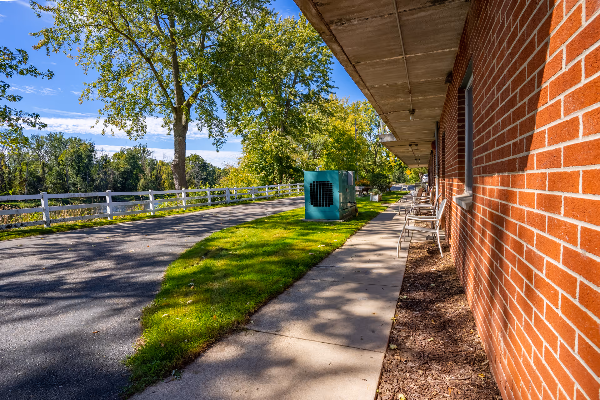 Sidewalk along a red brick building with several metal chairs lined up outside. A green utility box is visible on the grass next to the sidewalk. There is a paved road with a white wooden fence and tall trees with green foliage under a blue sky with some clouds.