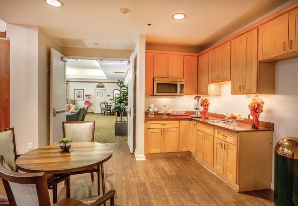 A small kitchen area with wooden cabinets, a microwave, a sink, and a countertop decorated with flowers and a fruit bowl. In the foreground, there is a round wooden table with two chairs and a small plant on the table. Through an open double door, a lounge area with chairs, a popcorn machine, and a dining table is visible.