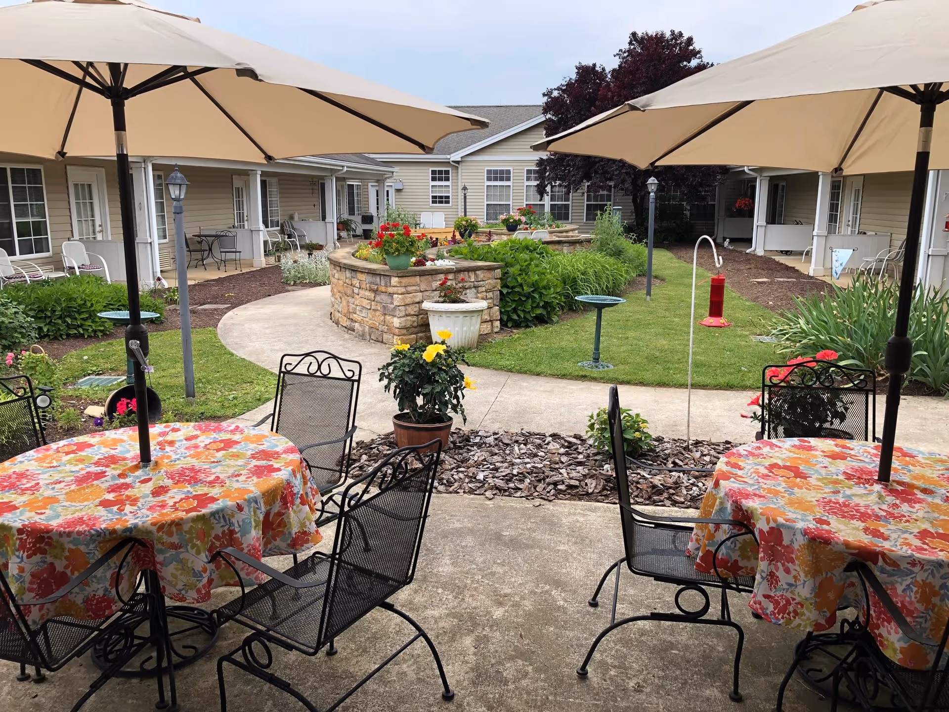 Outdoor patio area with two round tables covered in floral tablecloths and surrounded by black metal chairs. Large beige umbrellas provide shade over the tables. In the background, there is a curved stone planter filled with flowers and greenery, a concrete walkway, and a beige building with multiple windows and doors. Various plants and garden decorations are visible throughout the courtyard.