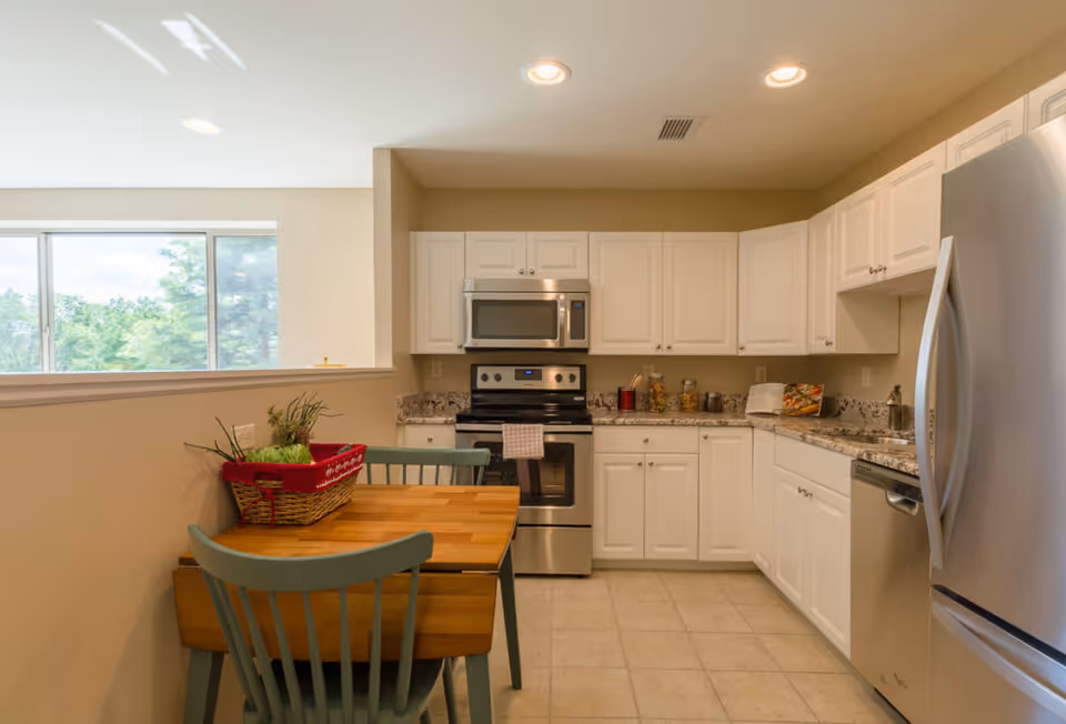 Well-lit kitchen with stainless steel appliances, white cabinets, granite countertops, and a small wooden dining table with green chairs.