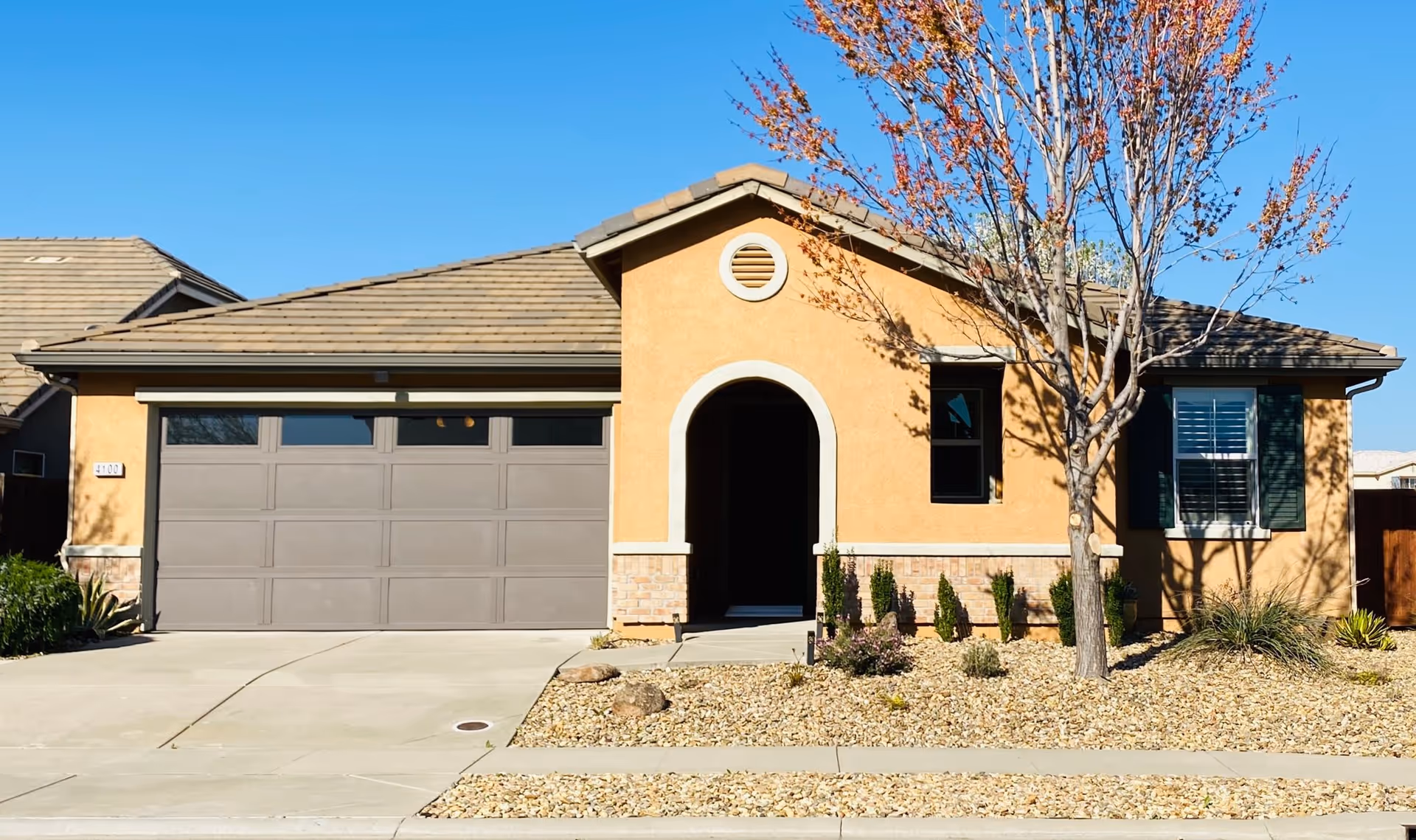 Single-story stucco house with arched entryway, two-car garage, and a front yard with gravel landscaping and a tree.