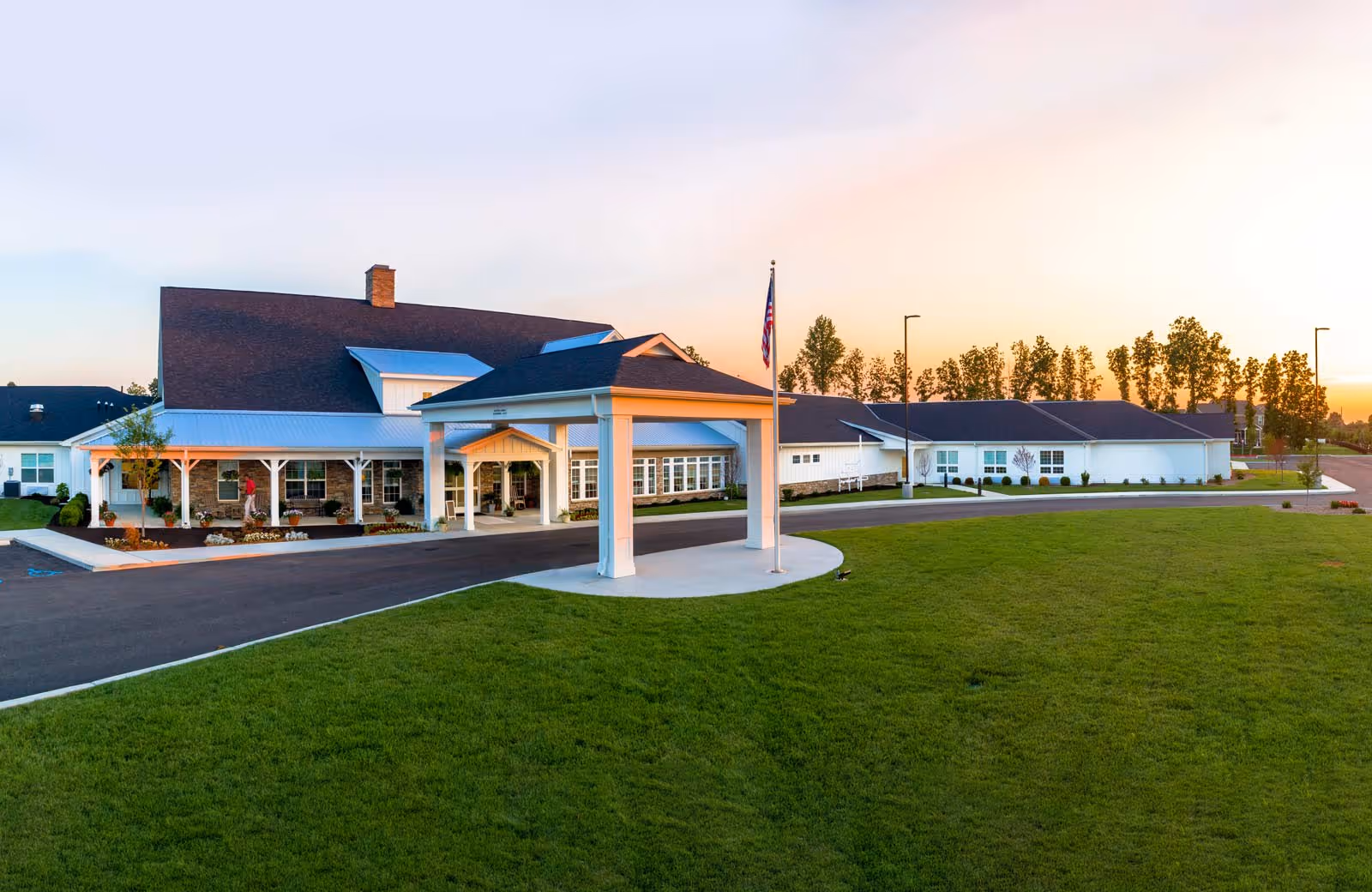 Exterior view of Demaree Crossing facility at sunset, showing a large building with a covered entrance, an American flag on a flagpole, a paved driveway, and a well-maintained green lawn in the foreground.