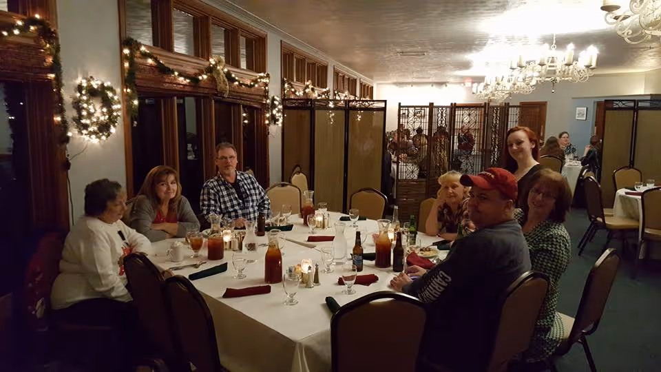 Several people seated around a decorated dining table in a warmly lit banquet room with wreaths and chandeliers.