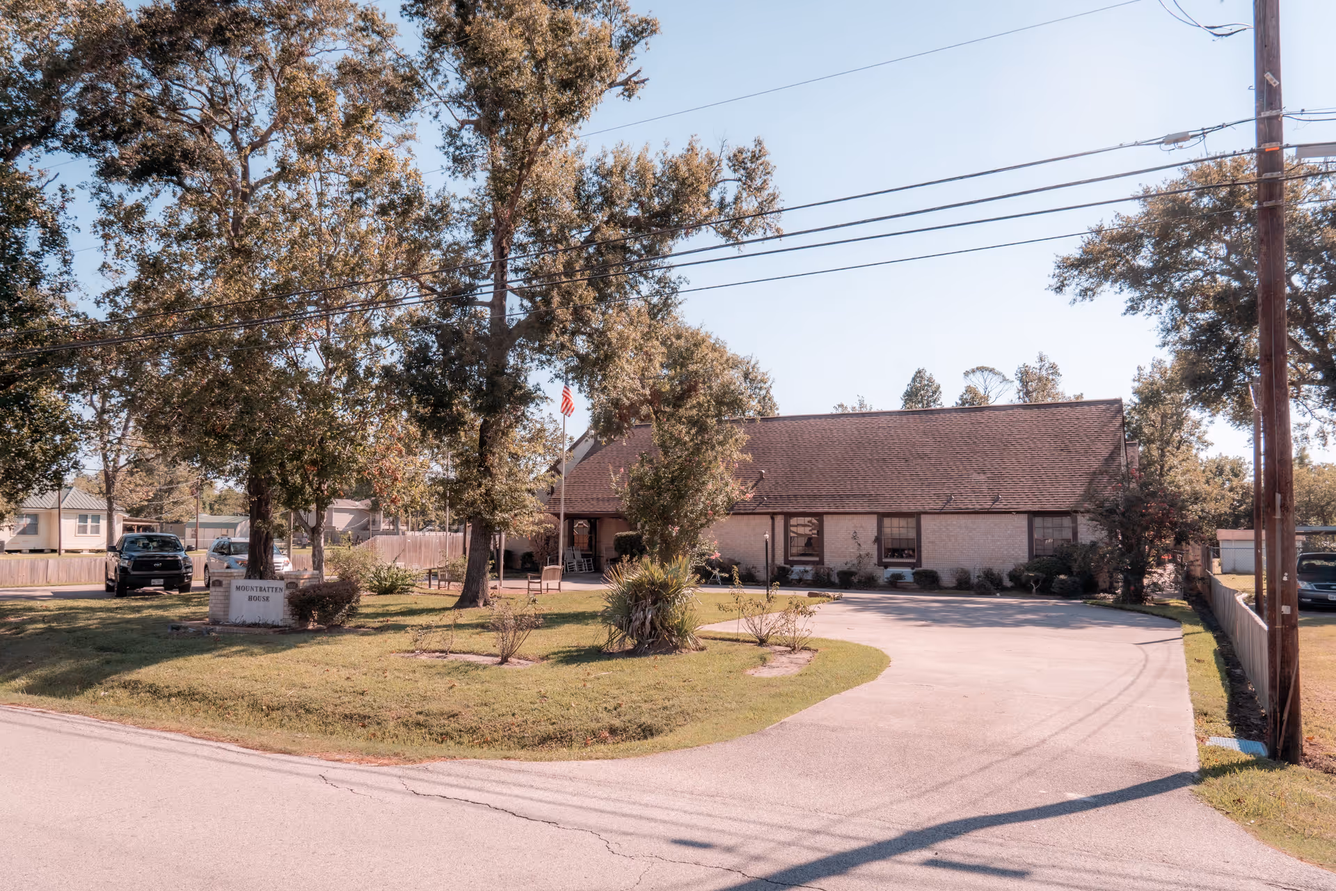 Single-story senior living building with a curved driveway, front lawn, trees, and an American flag flying near the entrance.