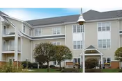 Three-story light-colored senior living building with a covered entrance, small trees and landscaped lawn in front.