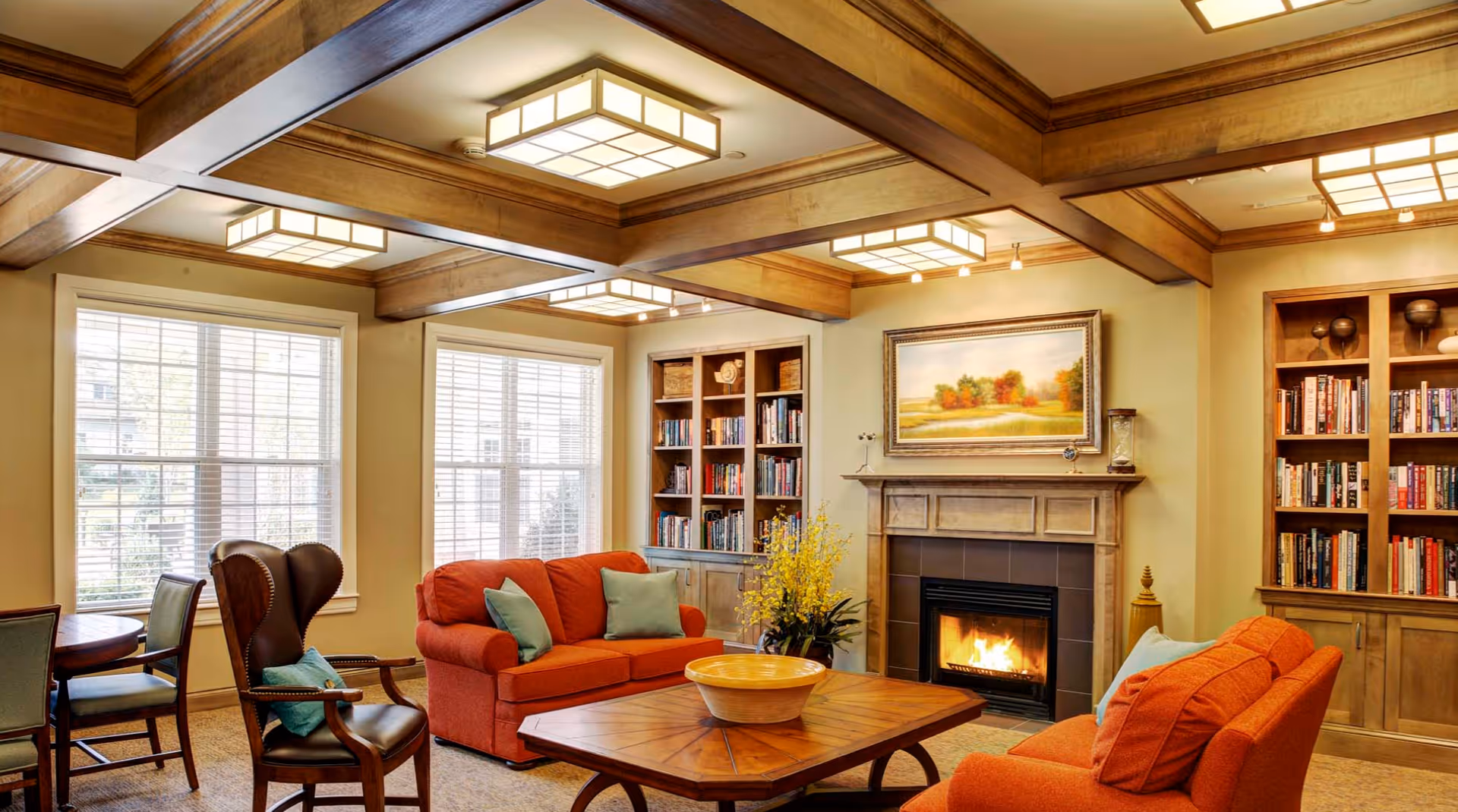A cozy living room with two orange sofas, a wooden coffee table with a decorative bowl, a leather armchair with a blue cushion, built-in bookshelves filled with books, a fireplace with a fire burning, and a painting of a landscape above the fireplace. The room has large windows with white blinds and wooden ceiling beams with modern square light fixtures.