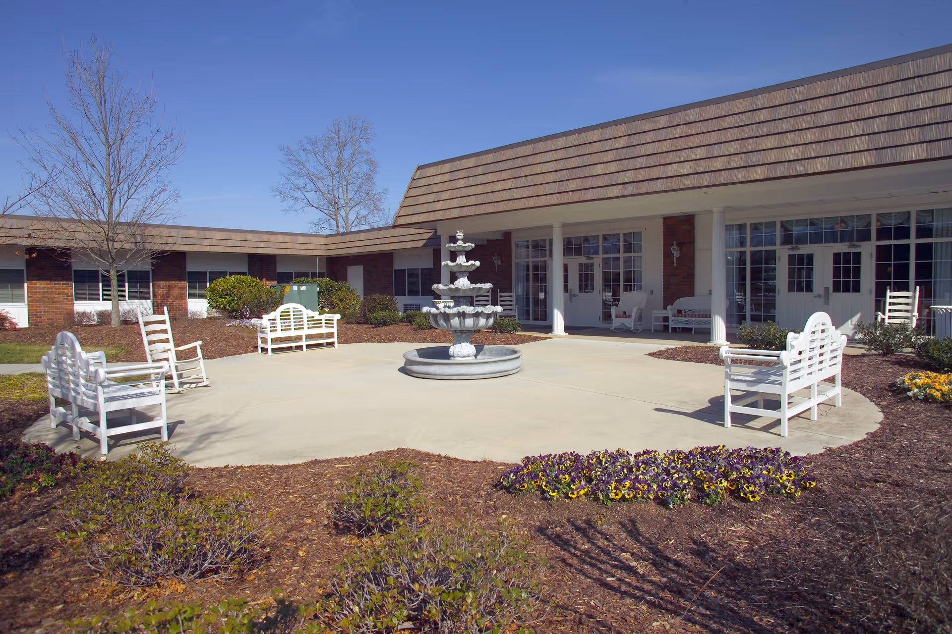 Outdoor courtyard area with a central multi-tiered fountain surrounded by white benches and rocking chairs. The courtyard is part of a single-story brick building with large windows and a shingled roof. There are some leafless trees and flower beds with purple and yellow flowers around the paved area.