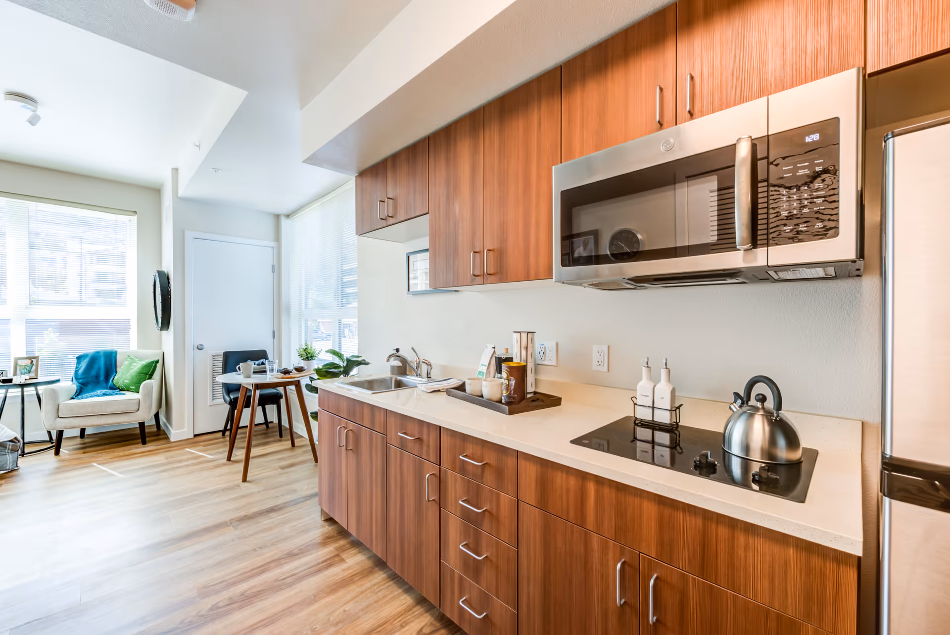 A modern kitchen area with wooden cabinets, a countertop with a sink, a stovetop with a kettle, and a microwave above. In the background, there is a small dining table with two chairs and a cozy armchair near large windows letting in natural light.