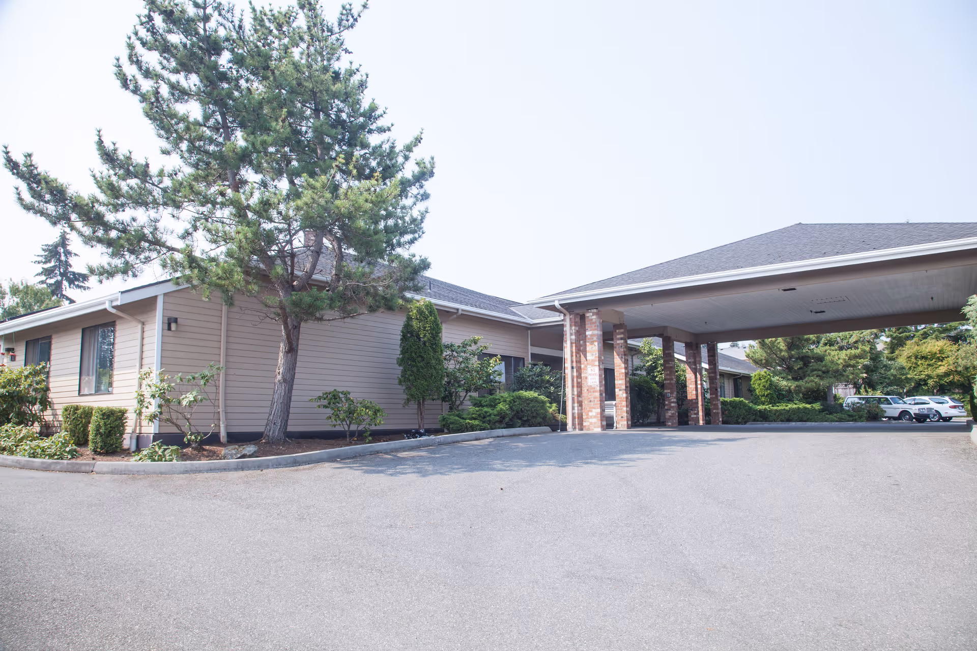 Exterior view of Cap Sante Court Retirement Community showing a single-story building with beige siding, a covered entrance supported by brick columns, surrounding greenery including trees and bushes, and a paved driveway with parked cars in the background.