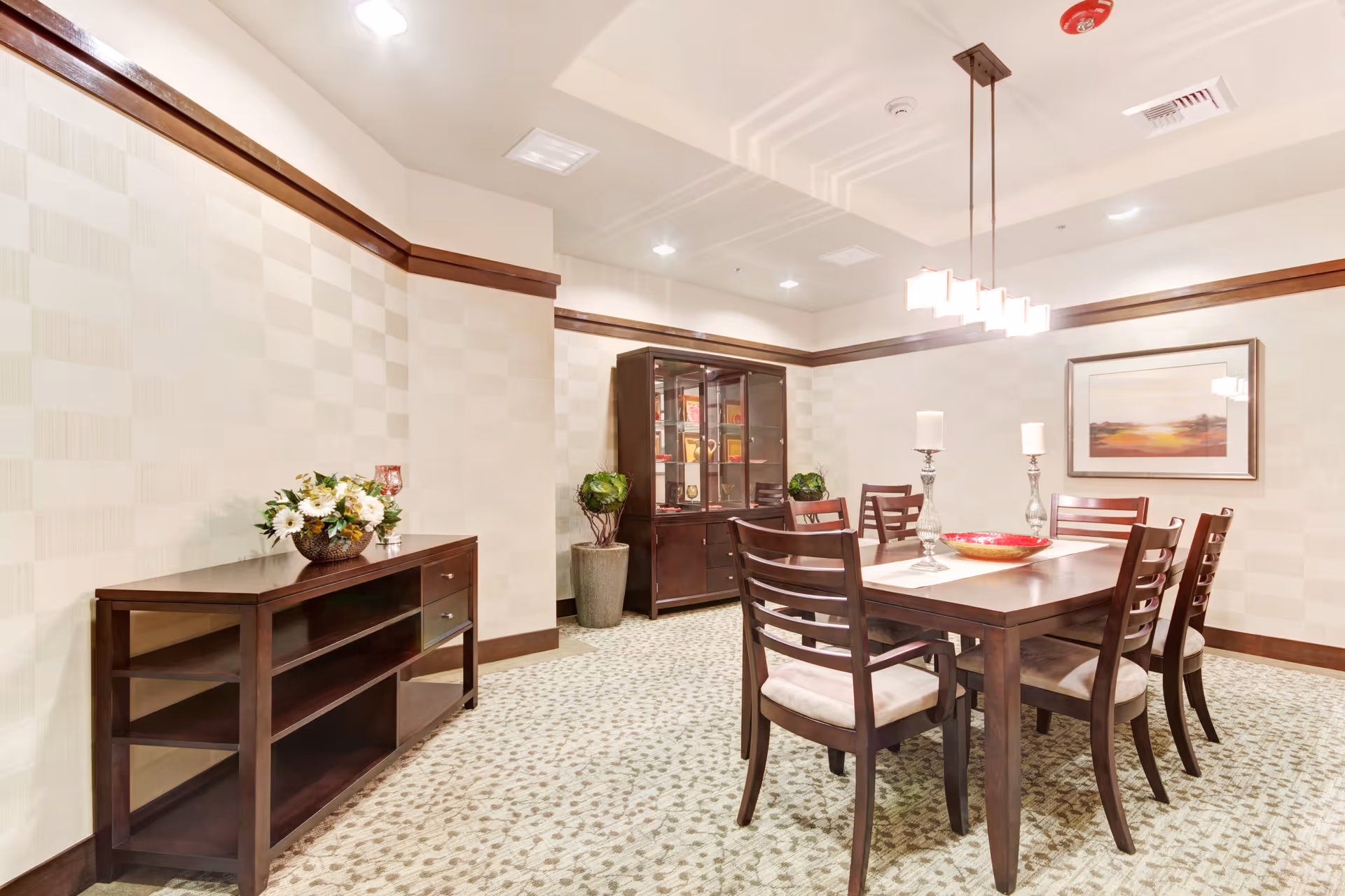 A well-lit dining room with a wooden dining table and six matching chairs with cushioned seats. The table is decorated with two tall candle holders and a decorative bowl. Against the wall is a wooden sideboard with a flower arrangement and a glass-front cabinet filled with books and decorative items. The walls have a subtle checkered pattern, and a framed landscape painting hangs on one wall. The room features recessed lighting and a modern rectangular chandelier above the table.