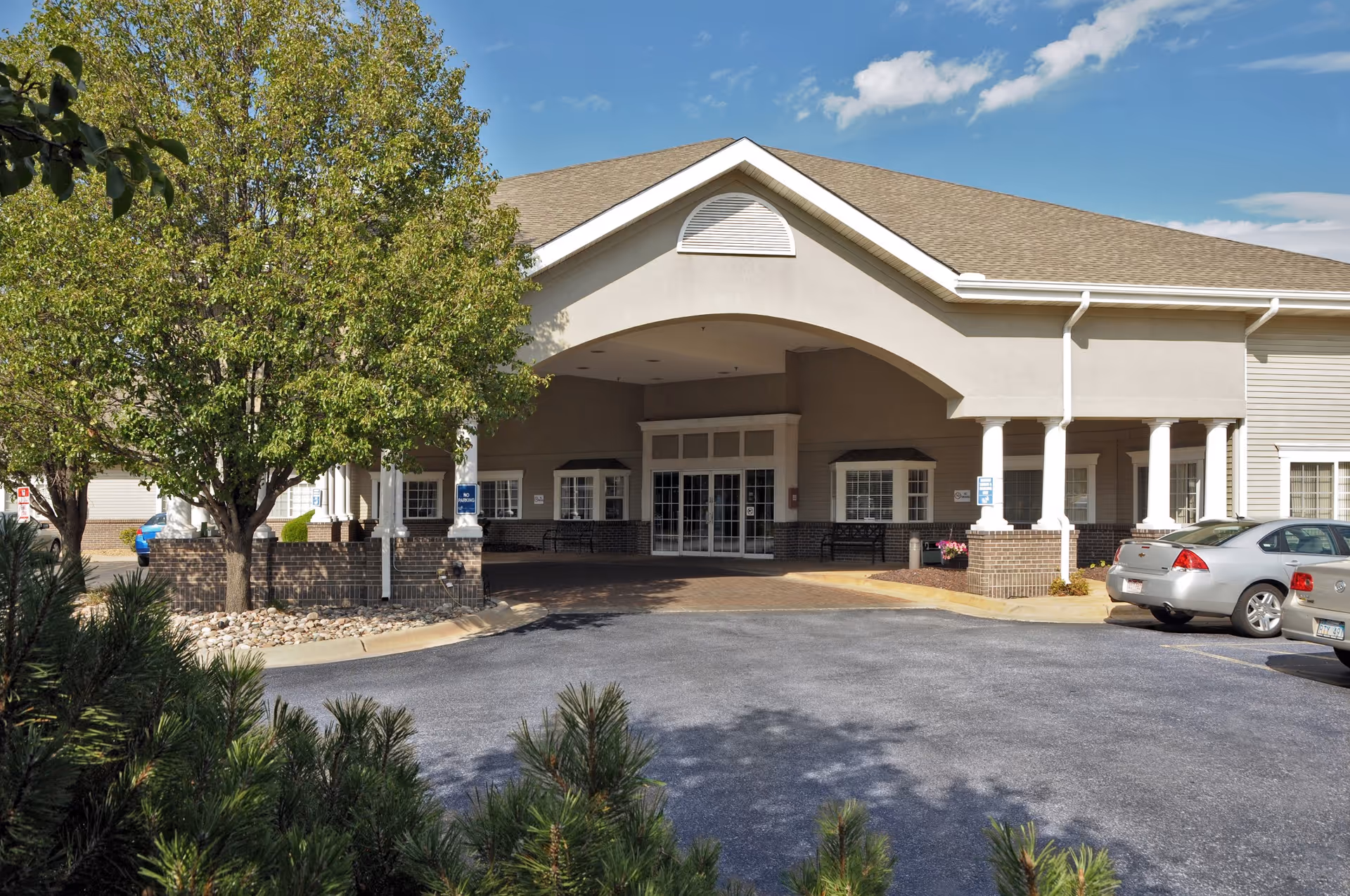 Front exterior view of a senior living facility building with a covered entrance, several parked cars, trees, and clear blue sky.