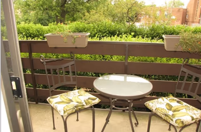 Small outdoor balcony with a round glass-top table and two metal chairs with cushions featuring a green leaf pattern. The balcony railing has two rectangular planters with green plants. In the background, there are trees and a building visible.