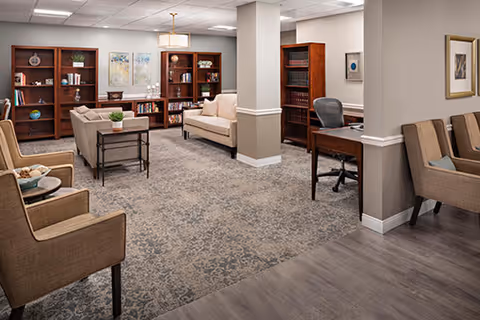 A spacious senior living facility common area with beige walls and carpeted floor. The room features multiple seating options including armchairs and sofas arranged around wooden bookshelves filled with books and decorative items. There is a desk with a chair in the corner and framed artwork on the walls. The lighting is provided by ceiling fixtures.