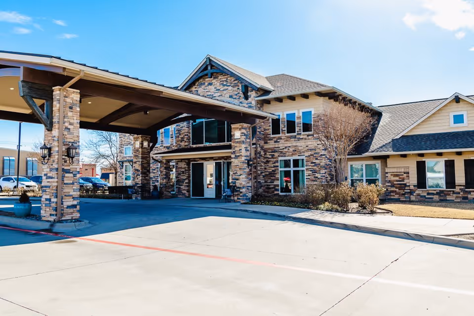 Exterior view of a senior living facility building with a covered entrance supported by brick pillars. The building features a combination of brick and siding with multiple windows and a clear blue sky above.