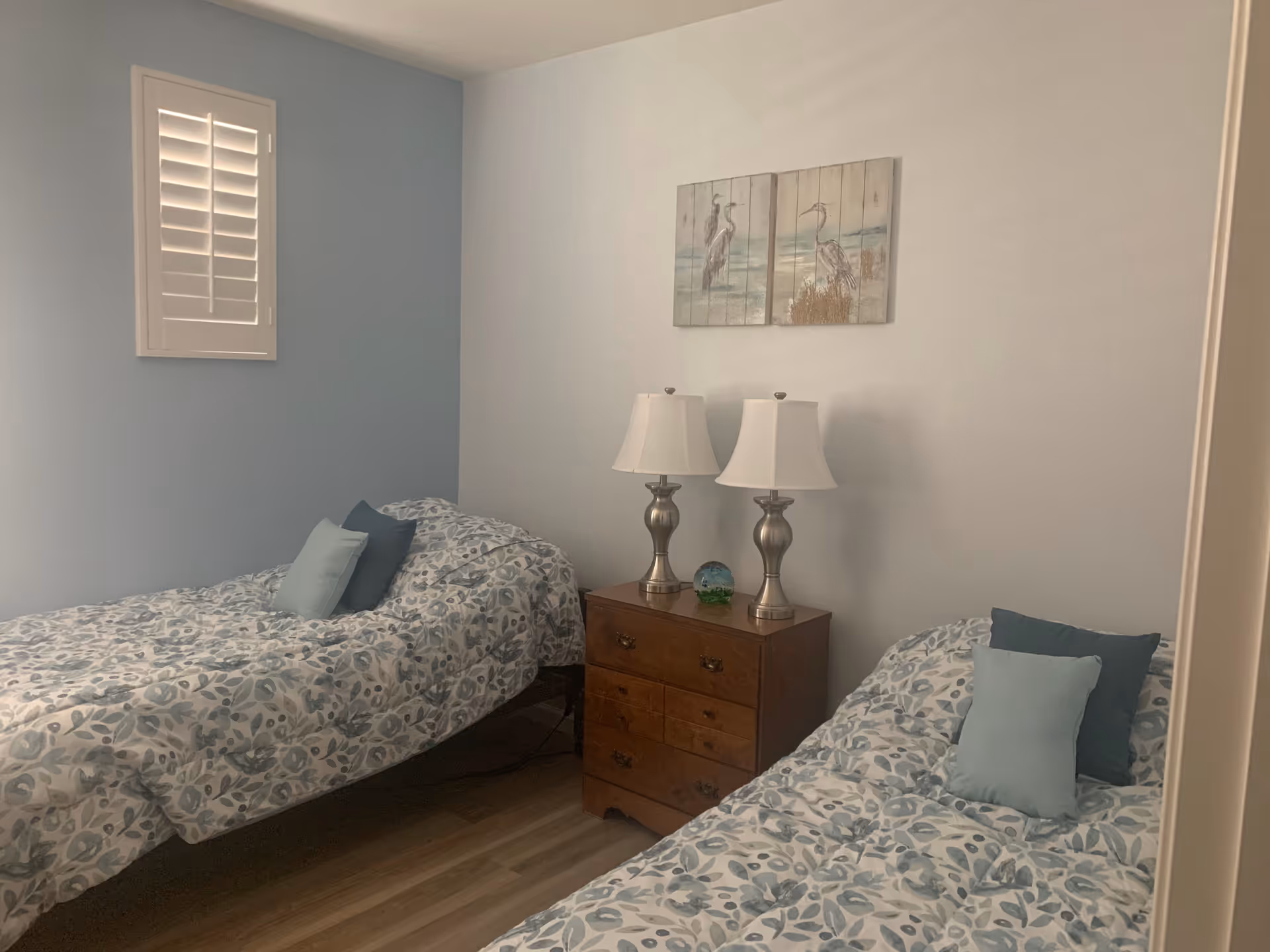 Small bedroom with two twin beds, patterned bedding, a wooden nightstand with two lamps, and coastal artwork on the wall.