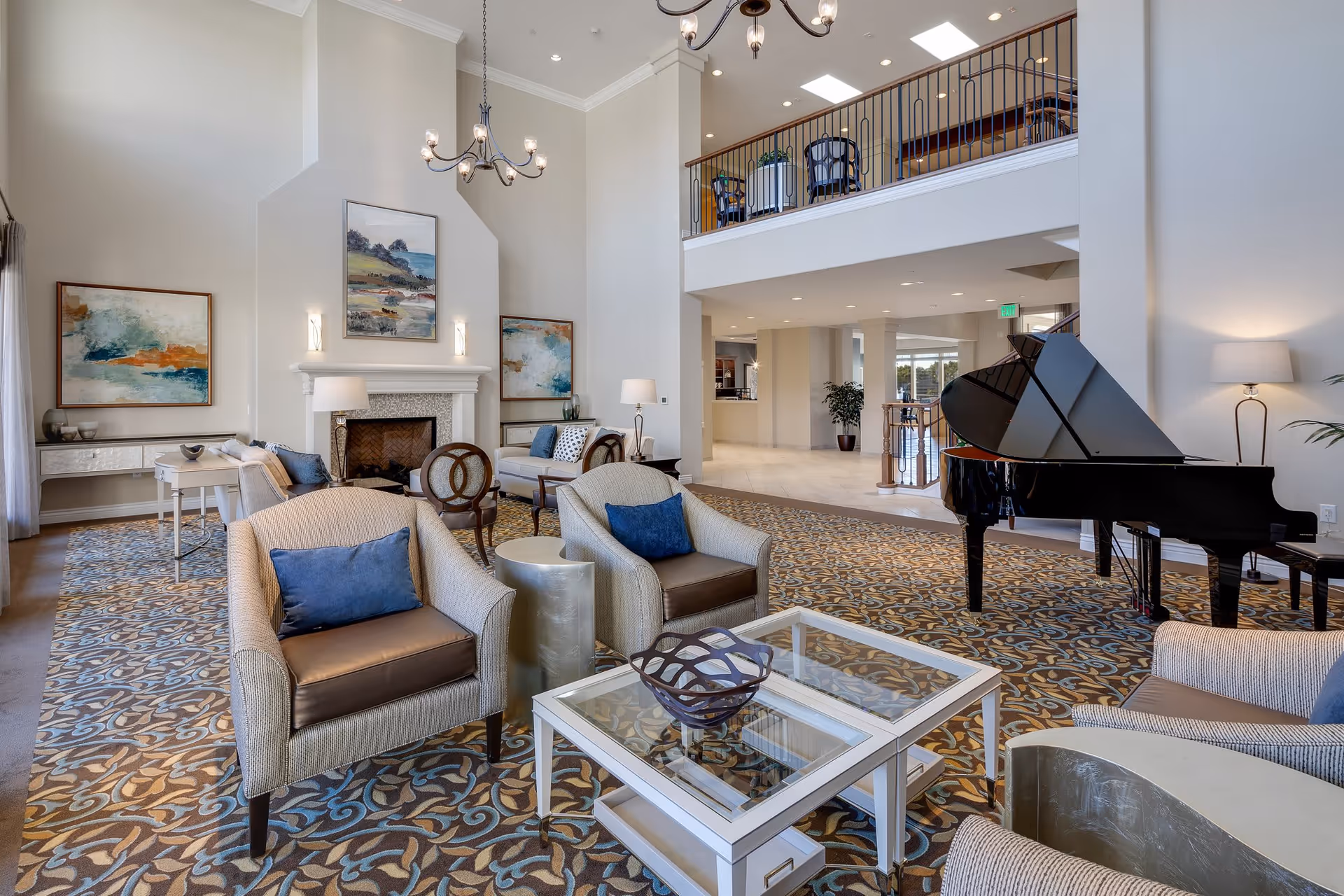 Spacious and elegant living room area with high ceilings, a grand piano, multiple armchairs with blue cushions, a glass coffee table, a fireplace with artwork above it, and a balcony overlooking the room.