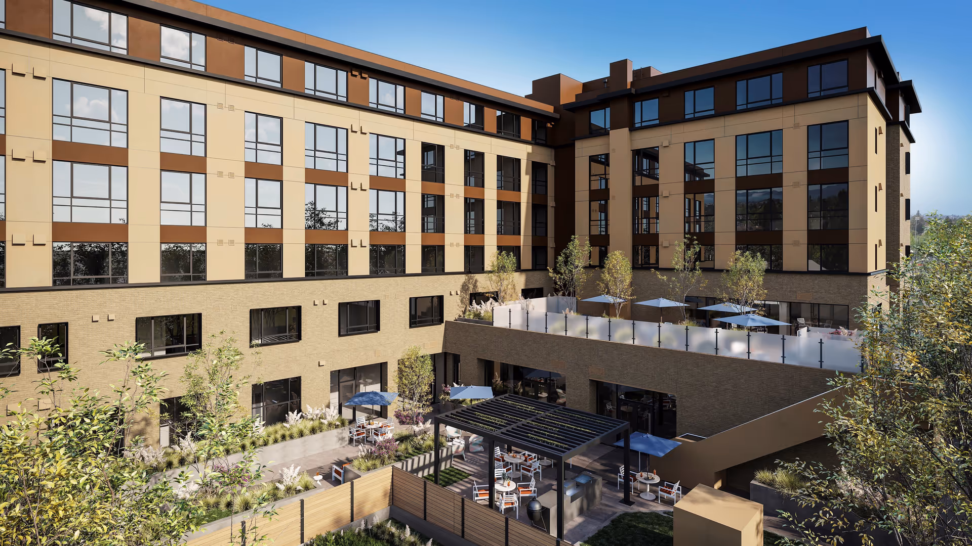 Outdoor courtyard area of a multi-story senior living facility with seating areas, tables with umbrellas, greenery, and a pergola, surrounded by the building's beige and brown exterior walls with large windows.
