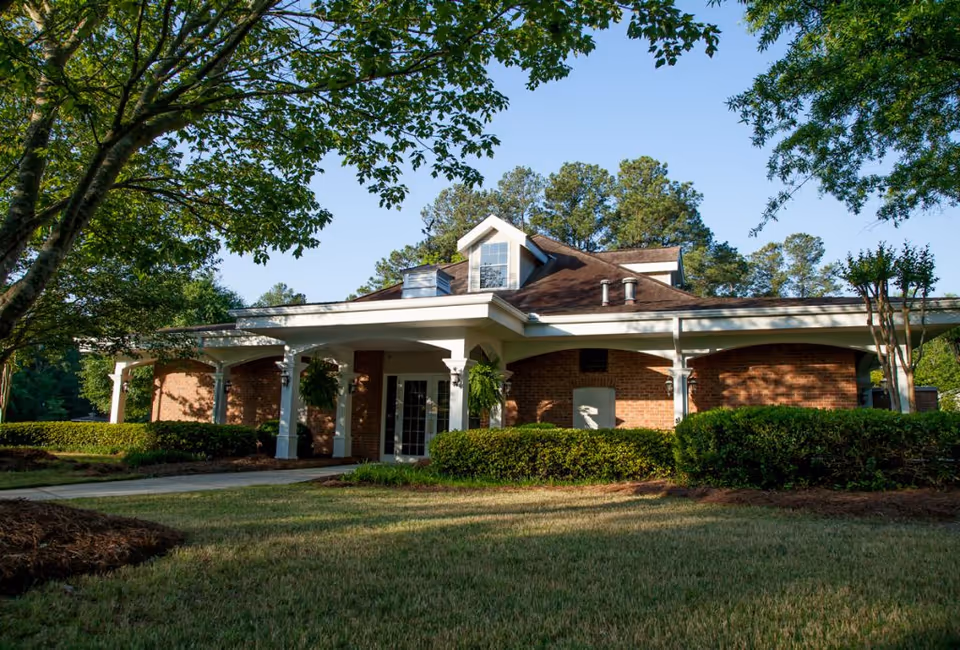 Exterior view of a single-story brick building with a covered entrance, surrounded by green bushes and trees under a clear blue sky.