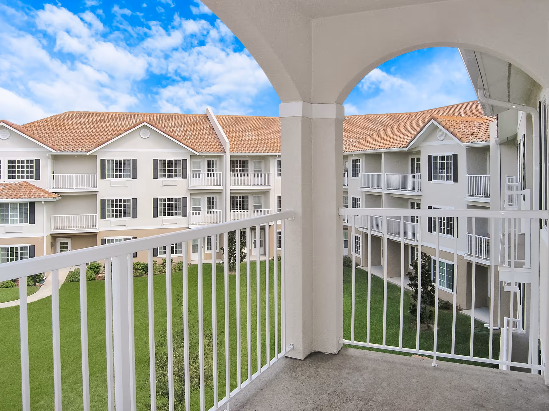 View from a balcony overlooking a courtyard with green grass and a three-story residential building with white railings, beige walls, and red-tiled roofs under a partly cloudy blue sky.