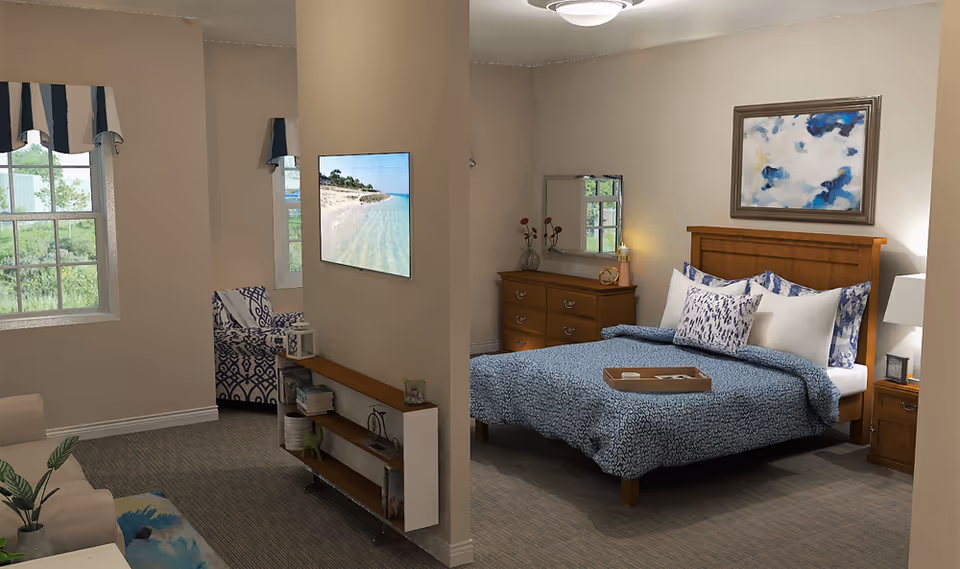 A cozy senior living facility bedroom with a wooden bed frame, blue patterned bedding, and multiple pillows. A wooden dresser with a mirror and decorative items is against the wall. A nightstand with a lamp and picture frame is beside the bed. The room has beige walls, carpeted floor, and a framed abstract painting above the bed. Adjacent to the bedroom is a small sitting area with a patterned armchair, a small shelf with books and decor, and two windows with blue and white valances letting in natural light.
