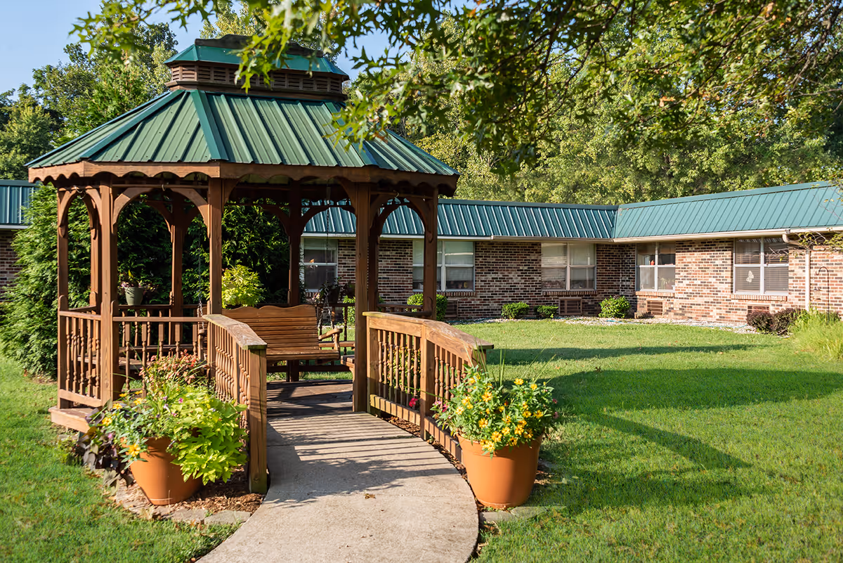Wooden gazebo with a green metal roof and potted flowers in a grassy courtyard in front of a single-story brick building.