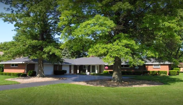 Single-story brick building with a gray roof surrounded by large green trees and a well-maintained lawn under a clear blue sky.