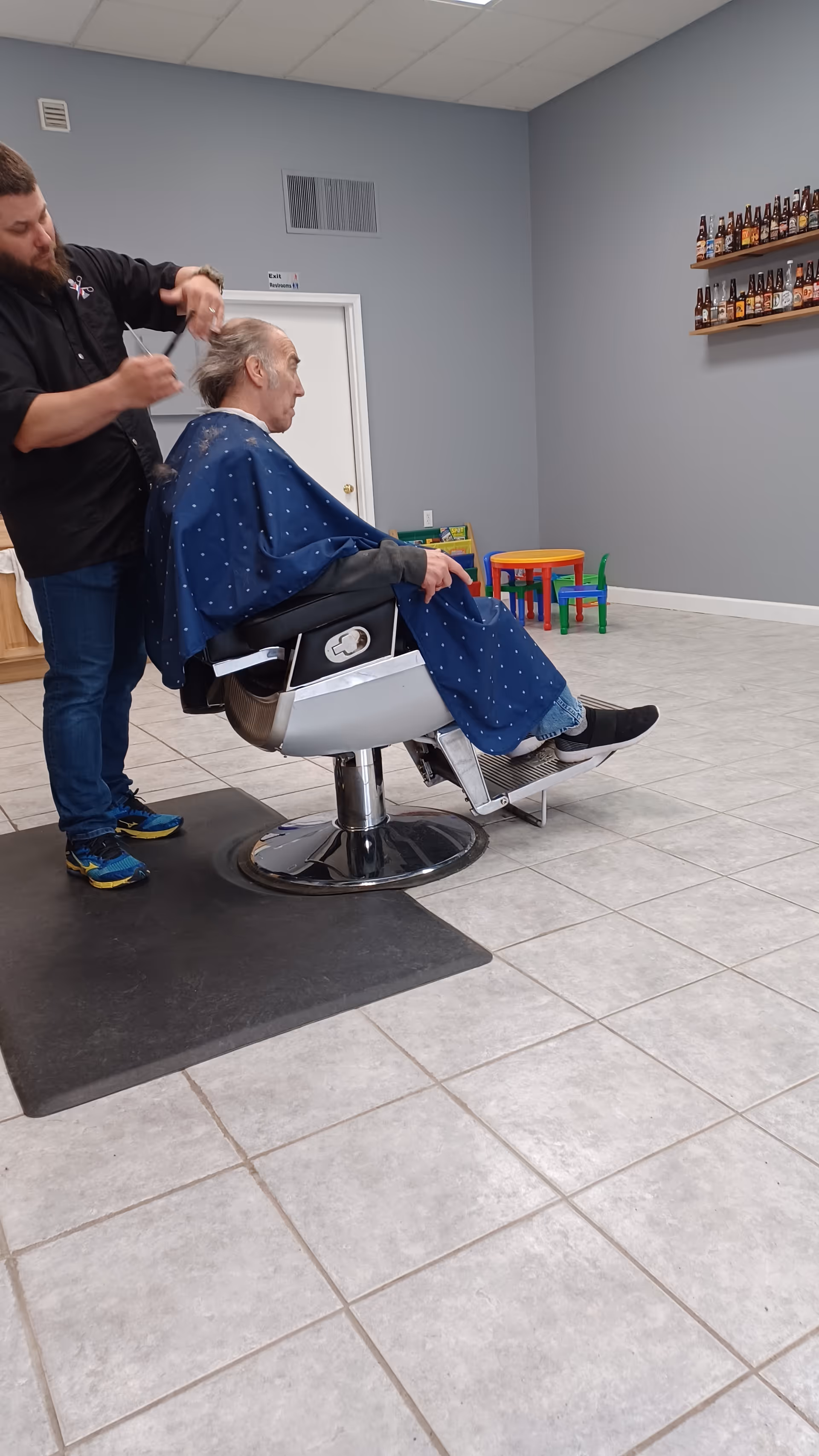 A man is getting a haircut from a barber in a room with tiled floors and gray walls. The man is seated in a barber chair covered with a blue cape, while the barber stands behind him cutting his hair. In the background, there is a small colorful children's table and chairs, and shelves mounted on the wall holding various bottles.