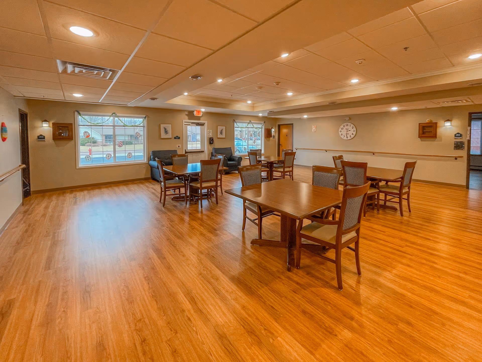 Spacious senior living dining/activity room with multiple wooden tables and chairs, lounge seating near windows, and wood-look flooring.