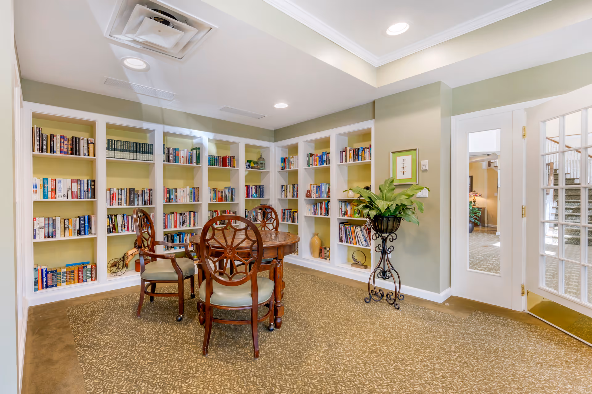 Bright communal library with built-in bookshelves, a round wooden table and chairs, and a potted plant by glass doors.