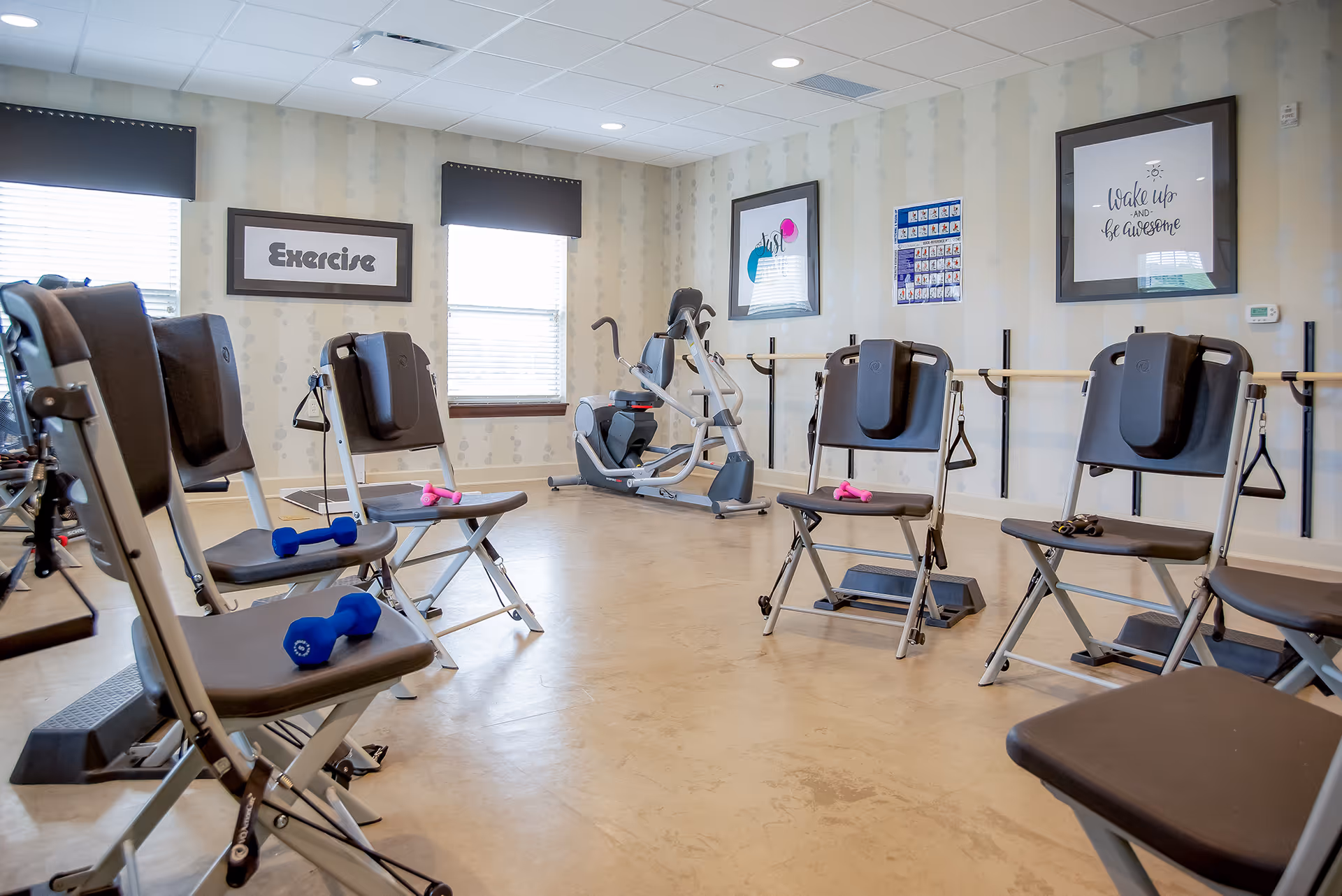 A bright exercise room with several folding chairs arranged in a circle, each equipped with small dumbbells and resistance bands. The room has light-colored walls with subtle patterns, two windows with black valances, and framed motivational posters on the walls. There is also an exercise machine in the corner and a ballet barre along one wall.