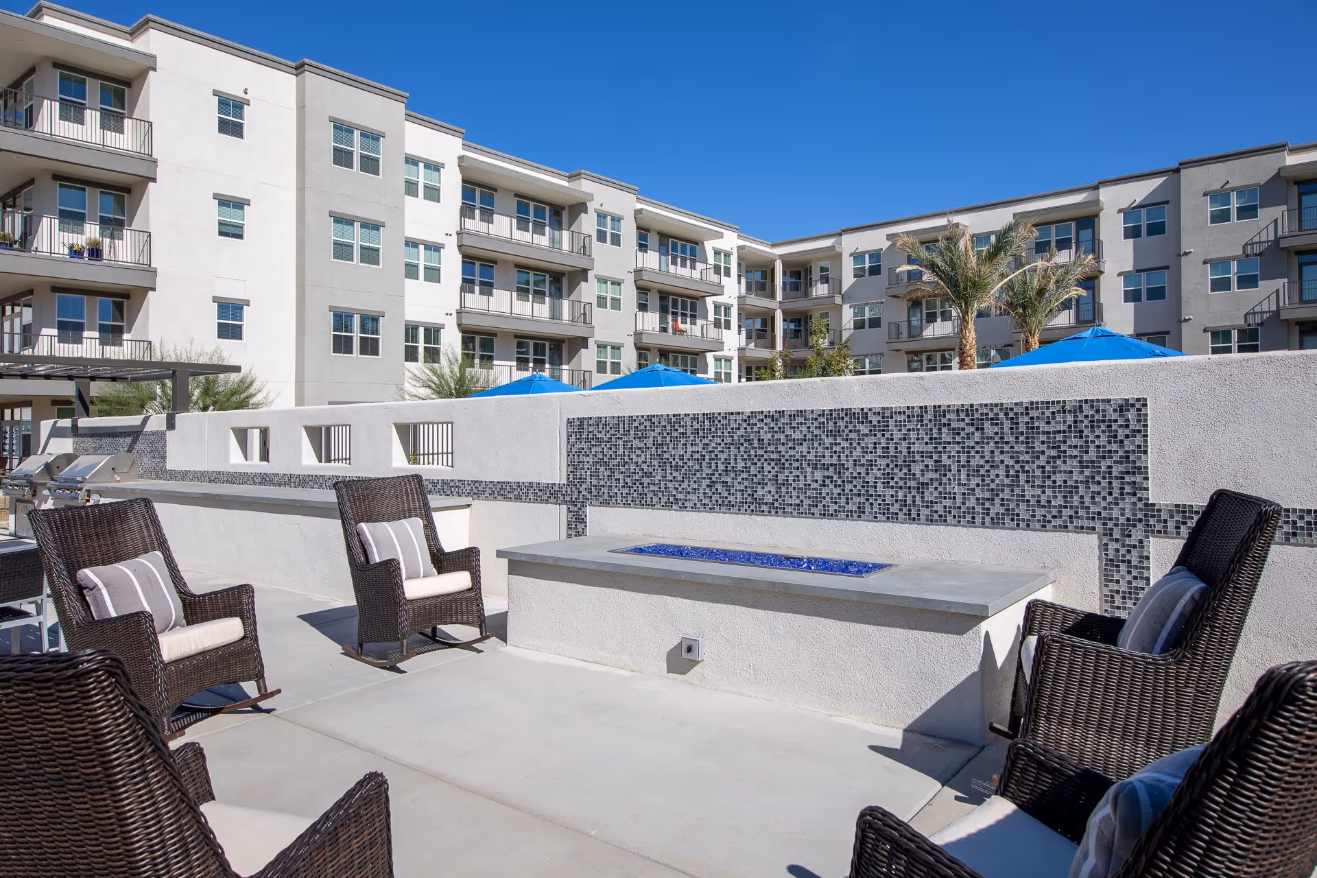 Outdoor patio area with wicker chairs and cushions arranged around a rectangular fire pit with blue glass stones, set against a white wall with decorative black and gray mosaic tiles. In the background, there are multi-story residential buildings with balconies, blue umbrellas, and palm trees under a clear blue sky.