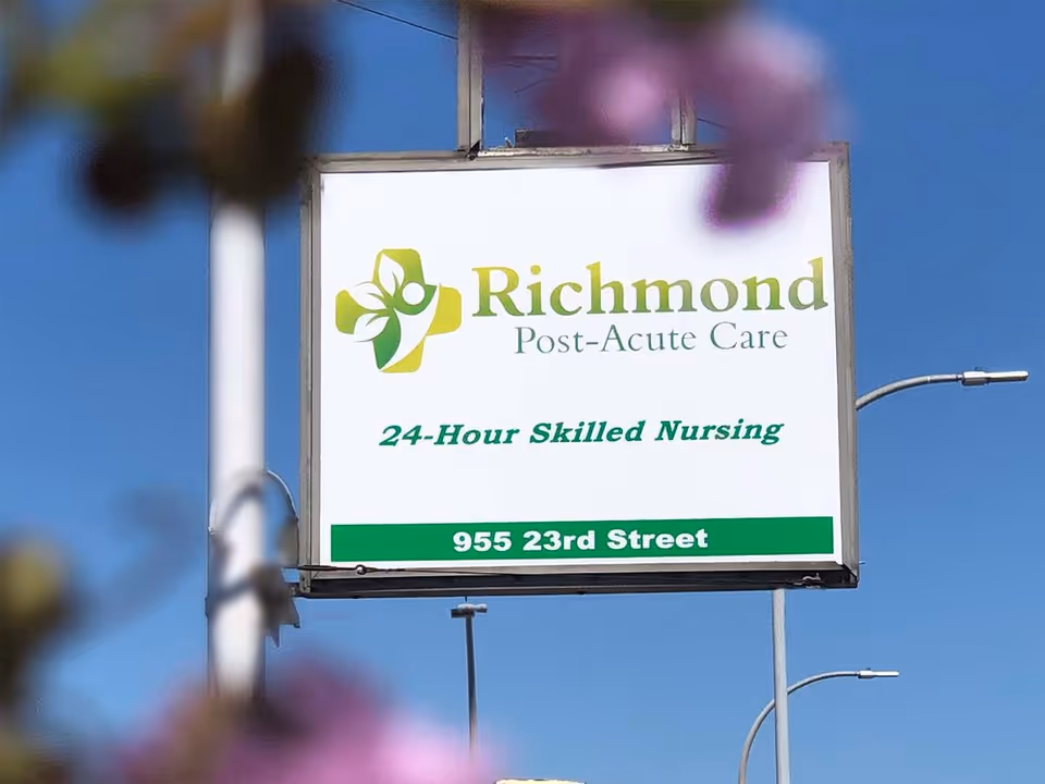 Outdoor sign for Richmond Post-Acute Care facility displaying their logo, the text '24-Hour Skilled Nursing', and the address '955 23rd Street' against a clear blue sky background.