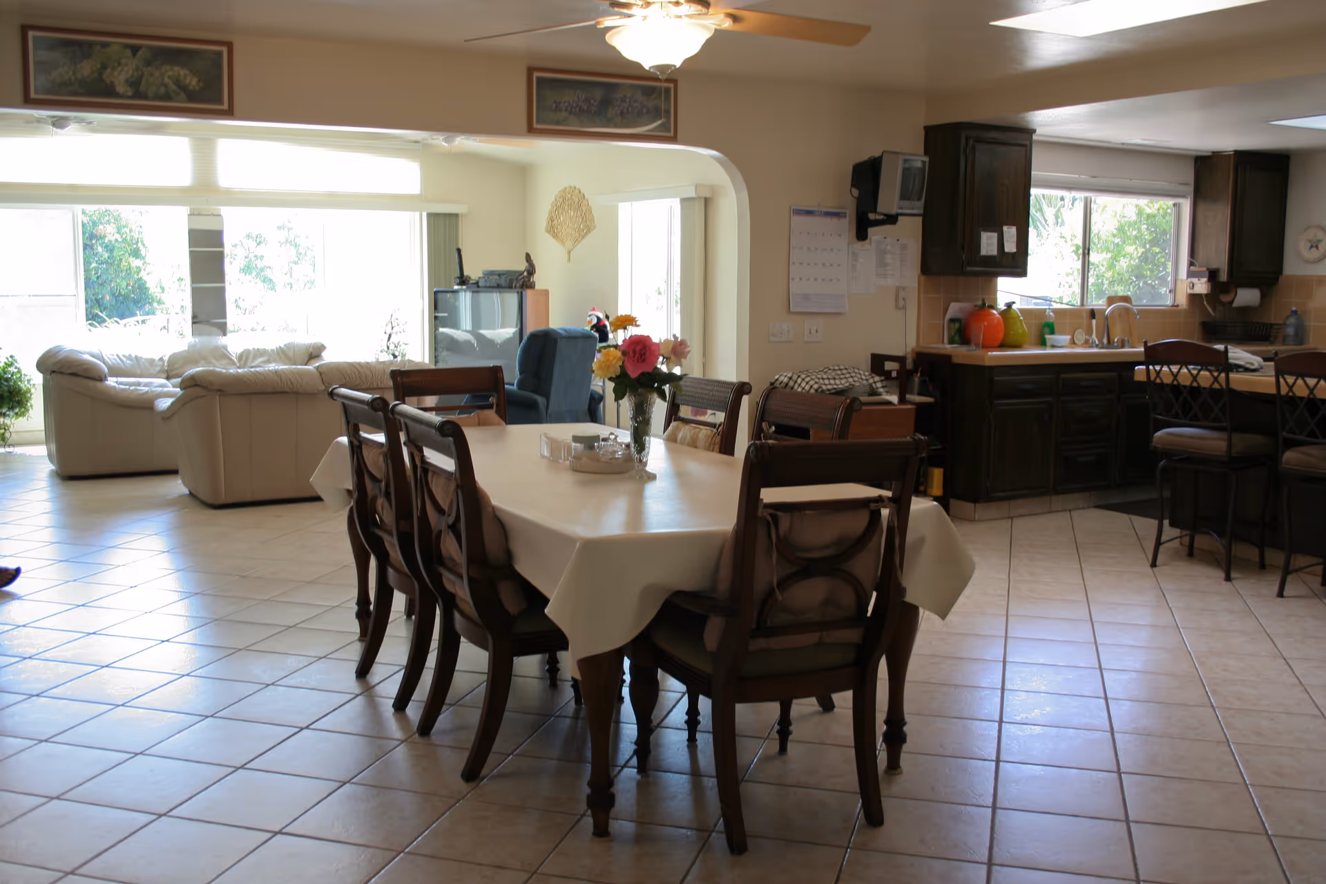A spacious open-plan room featuring a dining area with a rectangular table covered by a white tablecloth and surrounded by eight wooden chairs. A vase with colorful flowers is placed on the table. In the background, there is a living area with cream-colored sofas and a blue armchair near large windows letting in natural light. To the right, a kitchen area with dark wooden cabinets, a countertop, and bar stools is visible. The floor is tiled, and a ceiling fan with a light fixture is mounted above.