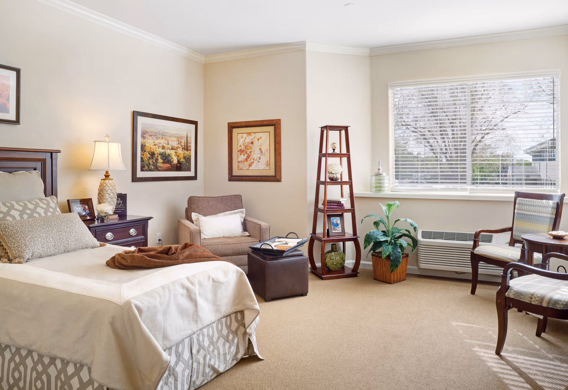 A bright, neatly furnished bedroom with a made bed, armchair and ottoman, shelving unit, and a large window.