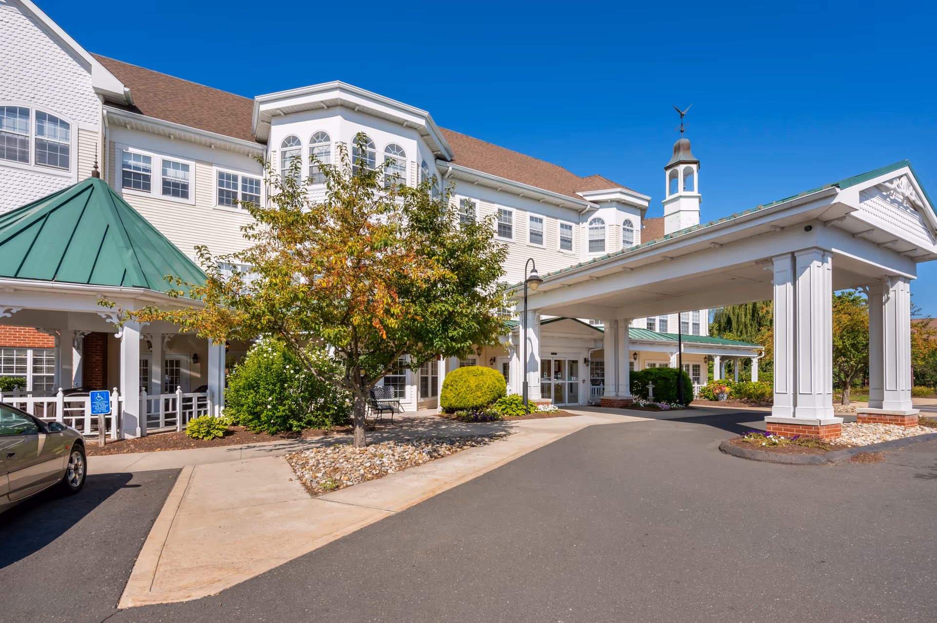 Exterior view of Brookdale South Windsor senior living facility on a clear sunny day. The building features white siding with multiple windows, a green metal roof over the entrance, and a covered drop-off area supported by white columns. There are trees and shrubs around the entrance and a paved driveway in front.