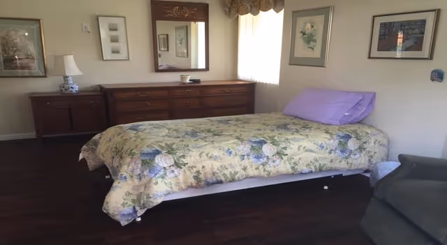 A bedroom with a single bed covered in a floral bedspread and a purple pillow. There is a wooden dresser with a mirror above it against the wall, a small lamp on a side table, and framed artwork hanging on the walls. A window with a valance lets in natural light, and a green armchair is partially visible in the corner.