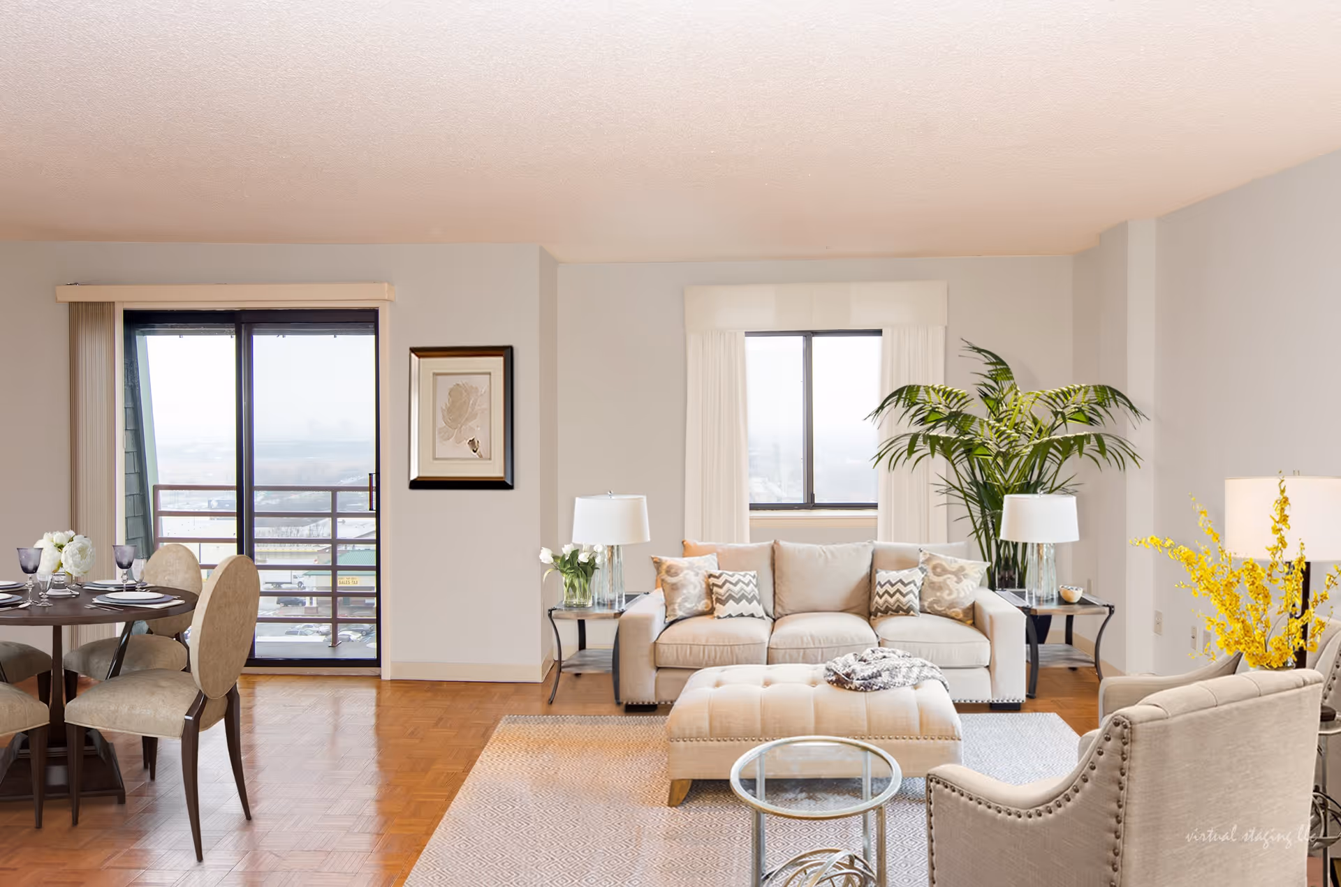 A bright and spacious living room with a beige sofa, matching armchair, and ottoman. The room features two side tables with lamps, a round glass coffee table, a large potted plant, and a dining area with a round table set for four near a sliding glass door leading to a balcony. The walls are light-colored with a framed artwork and windows letting in natural light.