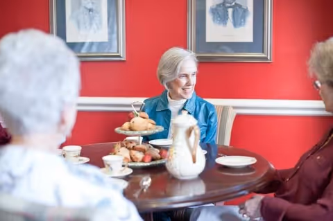 Three elderly women sitting around a round wooden table having tea and pastries in a room with red walls and framed portraits.