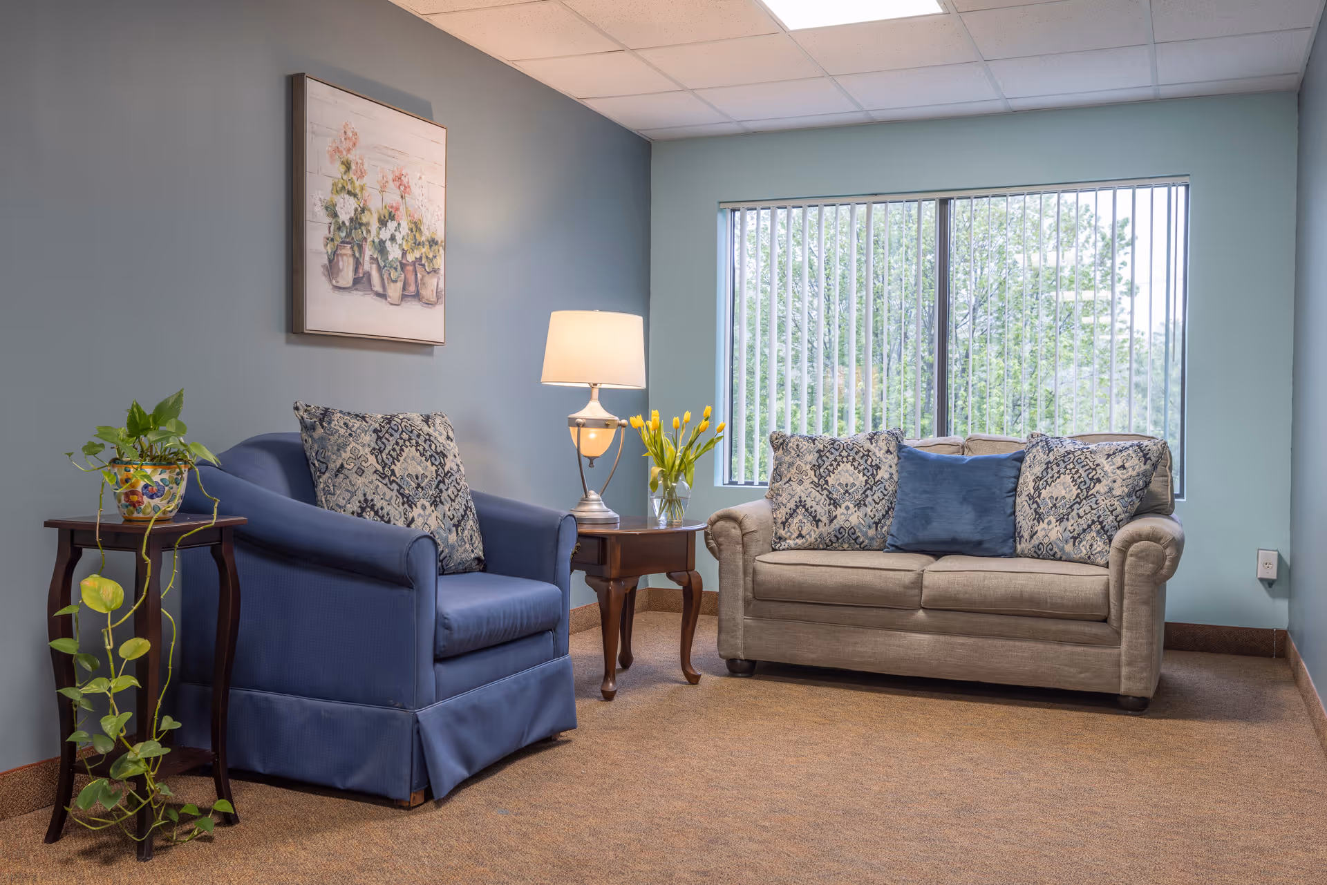 Cozy seating area with a blue armchair and beige loveseat, side tables with a lamp and plants, and a large window with vertical blinds.