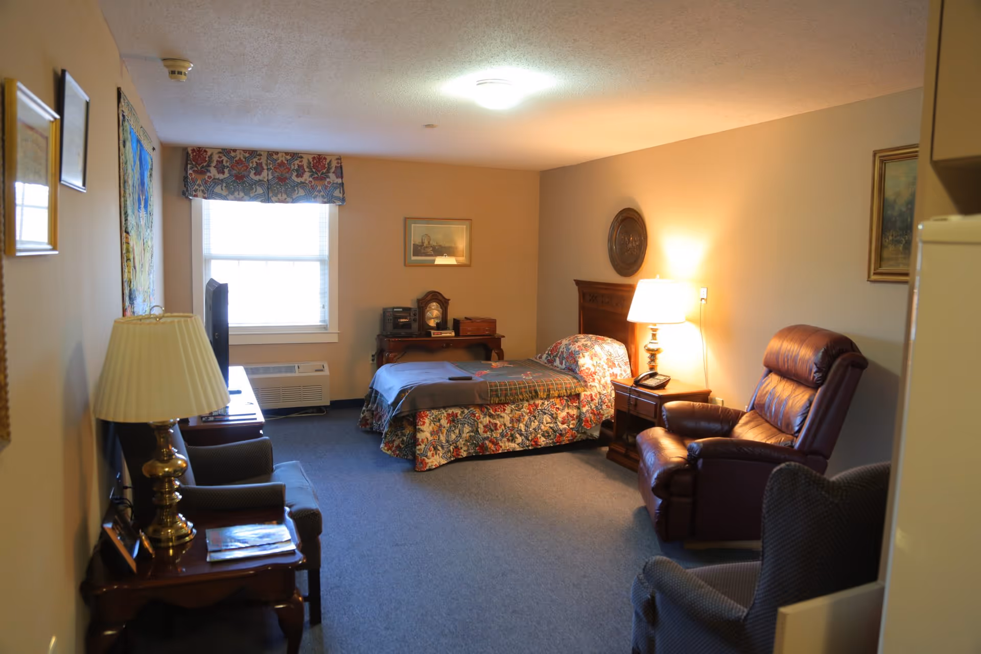 A cozy bedroom in a senior living facility with a single bed covered in a floral bedspread and a blanket. The room has a window with a floral valance, a wooden nightstand with a lamp and a telephone, a wooden desk with a clock and radio, a leather recliner, and two upholstered chairs. The walls are decorated with framed pictures and a decorative plate.