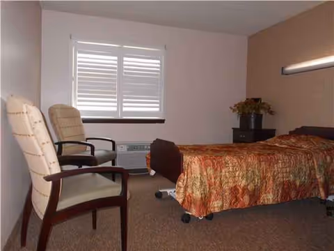 Senior living facility bedroom with a bed, two chairs, window blinds, and a nightstand.