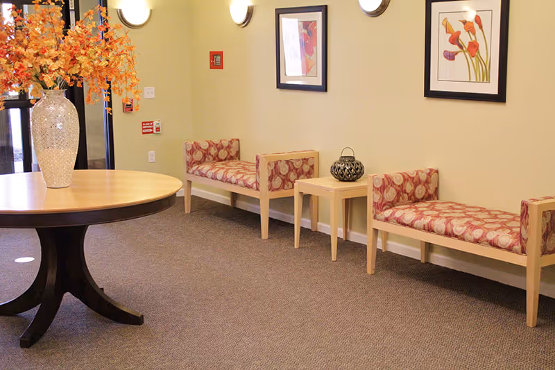 A senior living facility lobby with a round table holding a vase of orange flowers and patterned benches along a wall with framed artwork.